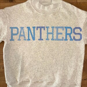 May include: A light gray heathered sweatshirt with a mock neck collar. The word PANTHERS is printed across the chest in large, block capital letters. Each letter features a distinct blue and white pattern, alternating between gingham and polka dots.