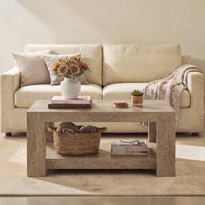 May include: A light-toned wooden coffee table with a lower shelf, featuring a textured white vase with sunflowers, a stack of books, a small potted succulent, and a woven basket containing folded textiles, positioned in front of a beige sofa.