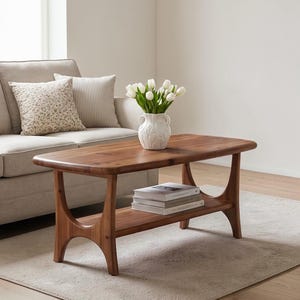 May include: A rectangular wooden coffee table with a smooth brown finish, featuring a lower shelf holding three stacked books. The table has curved, arched legs and supports a white ceramic vase filled with white tulips on its top surface.