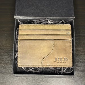 May include: A tan leather card holder with multiple horizontal slots and a curved stitched pocket detail. The brand name JILD LONDON is embossed in the bottom right corner. The item is presented inside a black box filled with black paper filler.