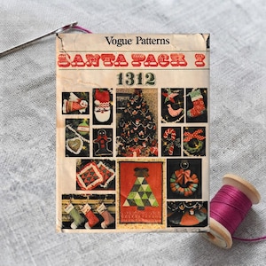 May include: A Vogue Patterns 1312 Santa Pack sewing pattern envelope featuring various Christmas-themed craft projects, including stockings, ornaments, a tree, and a wreath, displayed with a sewing needle and a spool of magenta thread.