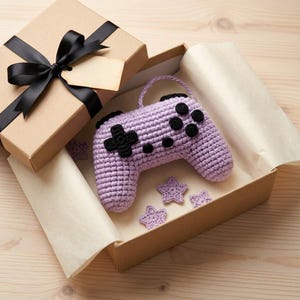 Pode incluir: Um controle de videogame de crochê em tom violeta claro com botões e direcional pretos, acompanhado por pequenas estrelas de crochê violetas, apresentado dentro de uma caixa de presente de papelão aberta com uma fita preta.