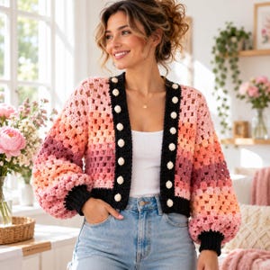 May include: A cropped crochet cardigan featuring a gradient of pink, peach, and orange yarn in a granny square pattern. The garment has long, voluminous sleeves with black ribbed cuffs, a black front placket with white buttons, and a black hem.
