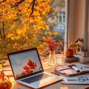 Op de afbeelding: Een zilveren laptop met een rood esdoornblad op het scherm, geplaatst op een houten bureau met een open notitieboek, een bril, een glazen vaas met droogbloemen, een kleine pompoen en een metalen pennenhouder bij een raam met herfstbomen.