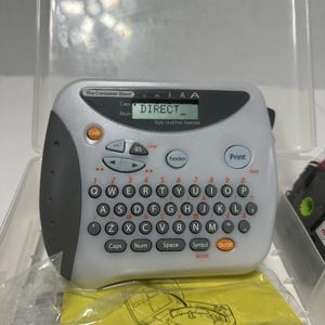 Puede incluir: Una rotuladora electrónica de color gris claro y gris oscuro con teclado QWERTY, una pantalla digital que muestra el texto DIRECT y un botón Print prominente. El dispositivo cuenta con teclas de función naranjas y grises en una caja de plástico.