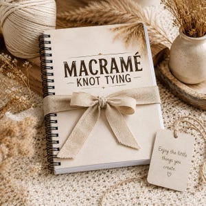Puede incluir: Un cuaderno de espiral con cubierta beige que presenta el texto MACRAMÉ KNOT TYING en marrón oscuro. Una cinta de tela de color tostado claro está atada en un lazo alrededor del centro. Una etiqueta rectangular con cordel dice Enjoy the little things you create.