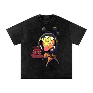 May include: A black acid-wash t-shirt featuring a graphic of a creature with a large circular head, sharp teeth, and red eyes, standing next to a small red pagoda-style structure. The creature has a yellow face and a brown, spotted body.