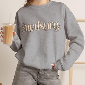 May include: A heather gray long-sleeve crewneck sweatshirt featuring the word "medsurg" printed in a cream-colored serif font across the chest, decorated with small floral illustrations in shades of pink, green, and yellow.