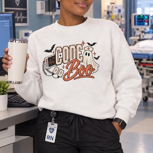 May include: A light gray crewneck sweatshirt featuring a graphic print with the text "CODE Boo" in orange and black. The design includes a ghost wearing a nurse cap, a medical heart monitor, and small bats. The sweatshirt has long sleeves.