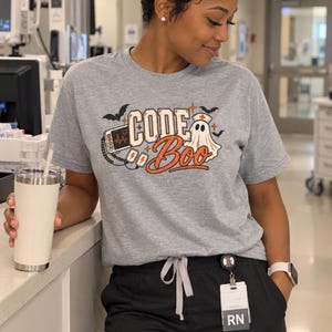 May include: A heather gray short-sleeve t-shirt featuring a graphic print with the text "CODE Boo". The design includes a medical heart monitor, a ghost wearing a nurse cap, bats, and stars in orange, black, and white colors on the front chest.