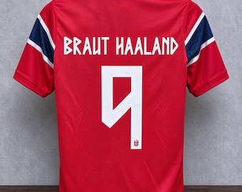 Puede incluir: Una camiseta deportiva roja de manga corta con el texto BRAUT HAALAND y el número 9 en blanco en la parte posterior. Las mangas tienen rayas azul oscuro y blancas. Un pequeño emblema de la bandera noruega se sitúa en la base del número 9.