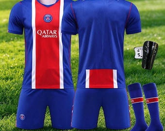 May include: A blue soccer jersey with a central red stripe and white accents, featuring the Qatar Airways logo and PSG crest. The set includes matching blue shorts, a pair of blue socks with a red stripe, and a black shin guard.