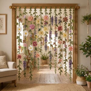 Op de afbeelding: Een decoratief deurgordijn met meerdere verticale strengen van gehaakte groene ranken versierd met diverse kleurrijke bloemen, waaronder witte madeliefjes, roze rozen en paarse lupinen, opgehangen aan een houten roede in een deuropening.