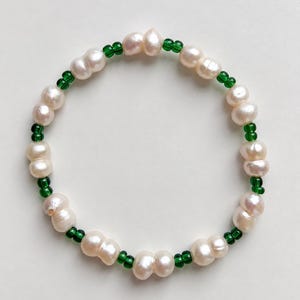 May include: A circular bracelet featuring alternating white freshwater pearls and small, translucent emerald green glass beads. The pearls have an irregular, organic shape, while the green beads are round and placed in pairs between each pearl.