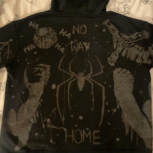 Puede incluir: Una sudadera negra con capucha que presenta un estampado gráfico gris desgastado en la espalda. El diseño incluye un emblema de araña central, el texto "NO WAY HOME", la palabra "HA" repetida, una calabaza, un brazo mecánico y una garra.