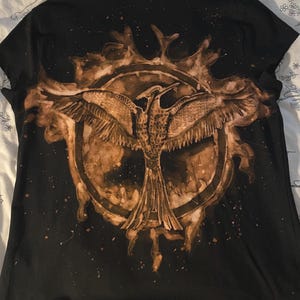 Könnte beinhalten: Ein schwarzes Kurzarm-T-Shirt mit einem gebleichten Motiv eines Vogels mit ausgebreiteten Flügeln, das in einem kreisförmigen Rahmen zentriert ist. Die Grafik hat ein verwaschenes, braun-beiges Aussehen mit spritzartigen Flecken.