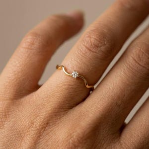 Può includere: Un sottile anello in oro caratterizzato da un delicato design a fascia ondulata. L'anello è incastonato con una singola piccola pietra preziosa rotonda e trasparente posizionata al centro dell'onda, per un aspetto sottile e raffinato.