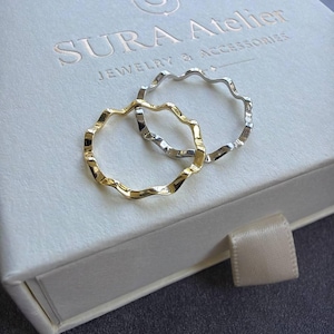May include: Two thin, wavy rings, one in a gold tone and one in a silver tone, resting on a white square box with the embossed text SURA Atelier JEWELRY & ACCESSORIES and a small ribbon pull tab on the front edge of the box lid.