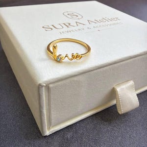 May include: A gold-toned ring featuring the word love in cursive script with a small clear stone set into the letter l. The ring rests on a white square jewelry box with the text SURA Atelier Jewelry & Accessories printed in dark brown ink.