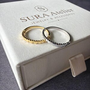 May include: Two thin rings, one gold-toned and one silver-toned, each featuring a continuous band of small heart shapes. The rings are resting on a white square box labeled with the text SURA ATELIER JEWELRY & ACCESSORIES and a logo.