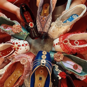 May include: A collection of eight ballet pointe shoes arranged in a circle, each uniquely decorated with lace, rhinestones, pearls, and floral embellishments in various colors including blue, red, white, black, and pink, featuring ornate center accents.