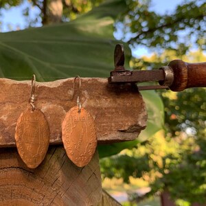 Op de afbeelding: Een paar koperkleurige, ovale oorbellen gemaakt van geperste munten, elk met een reliëfkaart van Illinois en de tekst Illinois. De oorbellen hangen aan zilverkleurige metalen haakjes en rusten tegen een ruwe stenen rand.