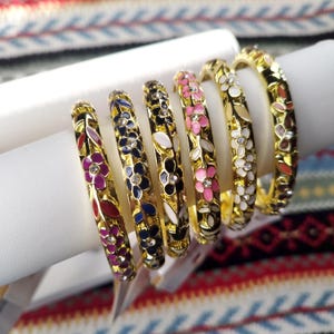 Peut inclure: Un ensemble de six bracelets rigides en métal doré, chacun présentant un motif floral répétitif rempli d'émail coloré dans des tons de rose, bleu, noir, blanc et rouge, avec de petits cristaux clairs incrustés au centre de chaque fleur.