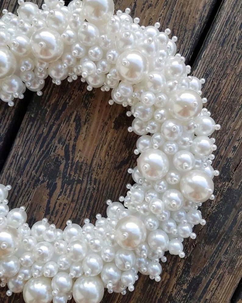 Puede incluir: Una corona de perlas blancas hecha de perlas de diferentes tama&ntilde;os. La corona est&aacute; sobre un fondo de madera marr&oacute;n.