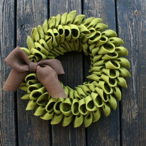 May include: A green burlap wreath with a brown burlap bow. The wreath is made of fabric loops and is perfect for a rustic or farmhouse decor style.