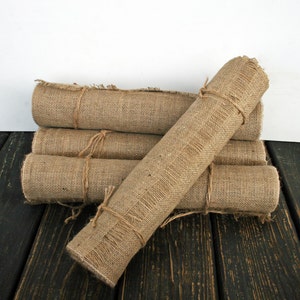 May include: Four rolls of natural burlap fabric, tied with twine, are stacked on a dark wood surface.