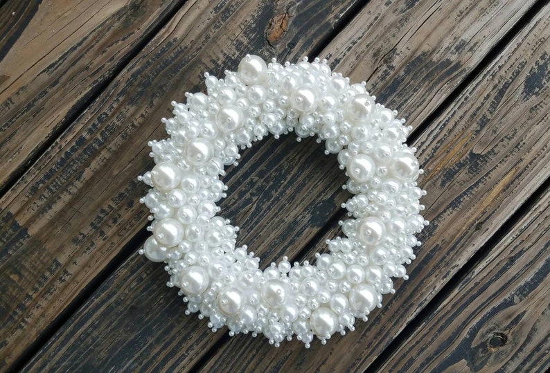 Puede incluir: Una corona de perlas blancas, hecha de muchas perlas peque&ntilde;as y grandes, dispuestas en forma circular. La corona est&aacute; sobre un fondo de madera marr&oacute;n.