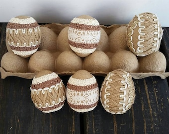 May include: Six brown Easter eggs decorated with white and brown lace and ribbon. The eggs are arranged in a cardboard egg carton.