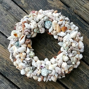 May include: A wreath made of various seashells in shades of white, brown, and beige. The wreath is arranged in a circular shape and is perfect for a coastal or beach-themed decor.