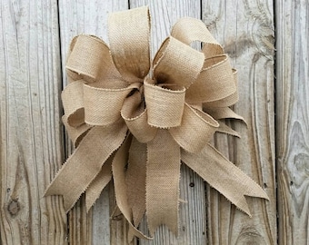 May include: A large, rustic burlap bow with a natural brown color. The bow is tied with a simple knot and has long, flowing tails.