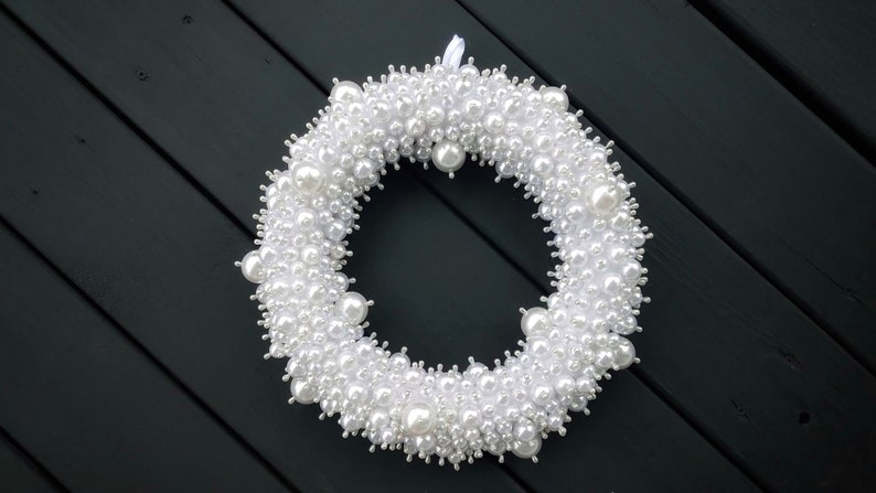 Puede incluir: Una corona de perlas blancas con una cinta blanca colgando de la parte superior. La corona est&aacute; hecha de muchas peque&ntilde;as perlas blancas y est&aacute; colgada sobre un fondo de madera negro.