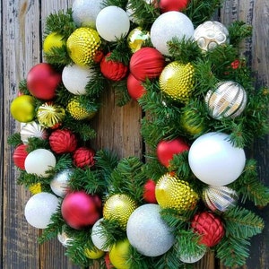 May include: A Christmas wreath decorated with red, white, gold, and silver ornaments. The wreath is made of green pine branches and is hanging on a wooden door.