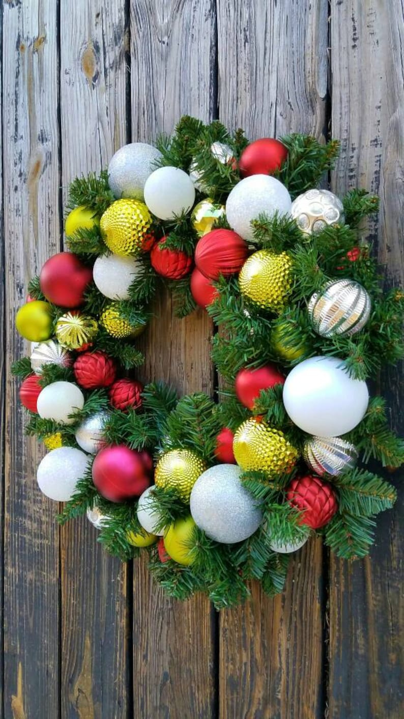 May include: A Christmas wreath decorated with red, white, gold, and silver ornaments. The wreath is made of green pine branches and is hanging on a wooden door.