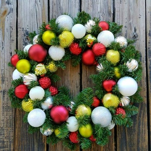 May include: A Christmas wreath decorated with red, white, silver, and gold ornaments. The wreath is made of green pine branches and is hanging on a wooden door.