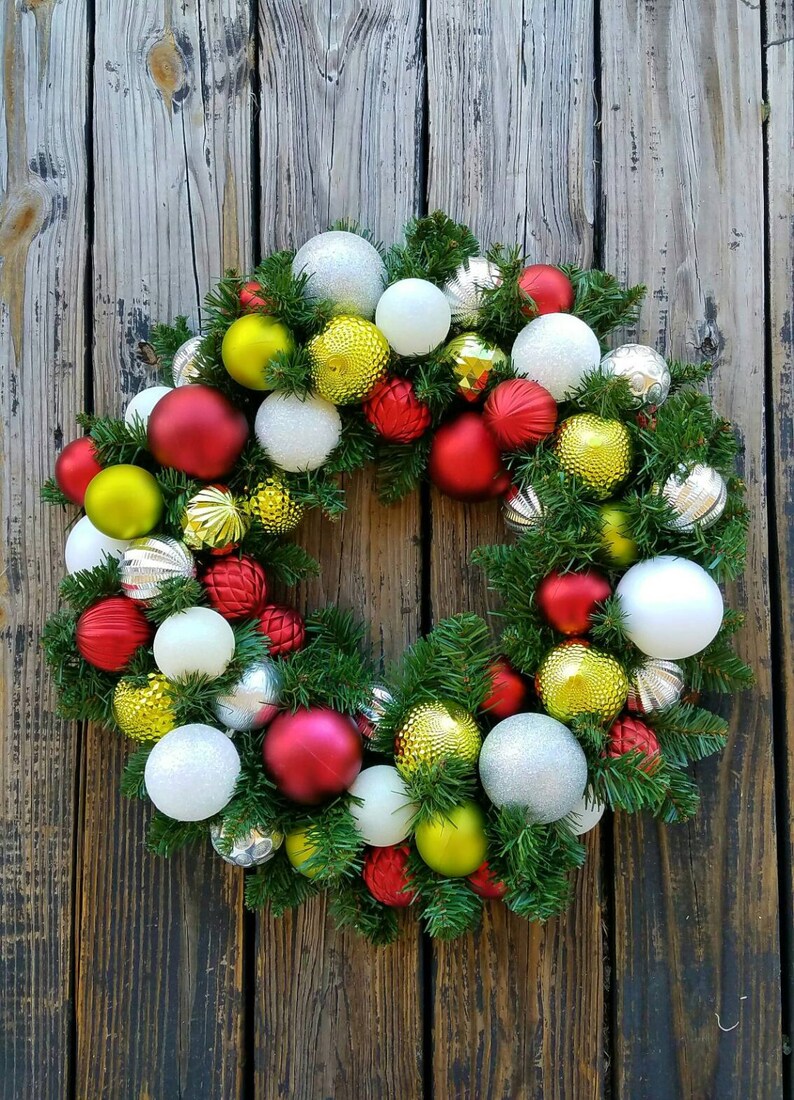 May include: A Christmas wreath decorated with red, white, silver, and gold ornaments. The wreath is made of green pine branches and is hanging on a wooden door.