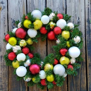 May include: A Christmas wreath decorated with red, white, green, and gold ornaments. The wreath is made of evergreen branches and is hanging on a wooden door.