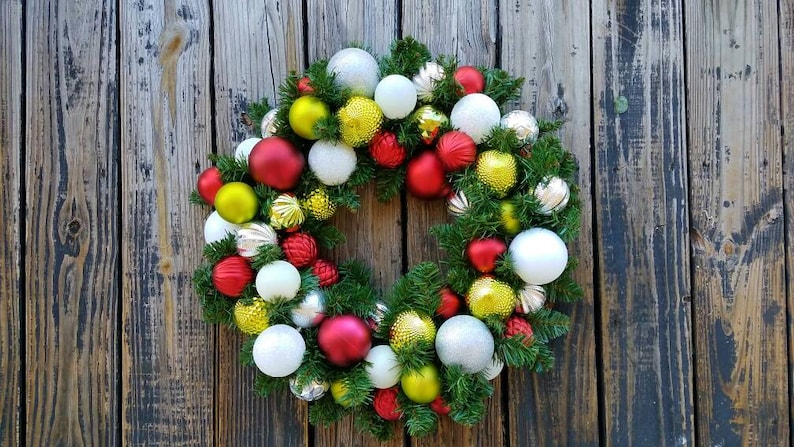 May include: A Christmas wreath decorated with red, white, green, and gold ornaments. The wreath is made of evergreen branches and is hanging on a wooden door.