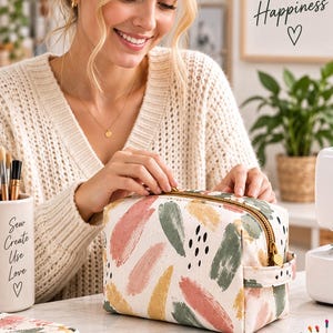 Puede incluir: Un estuche rectangular de tela con cremallera que presenta un fondo crema con un patrón de pinceladas multicolores en tonos rosa, verde salvia y amarillo mostaza, acentuado por pequeños puntos negros y una cremallera dorada.
