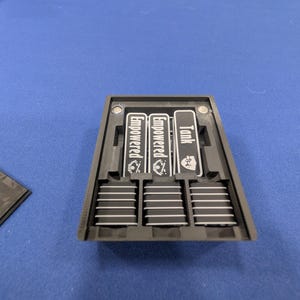 May include: A black rectangular plastic organizer tray containing three vertical slots with labels and three stacks of black tokens with white stripes. The labels read Empowered, Empowered, and Tank, each featuring a small white icon graphic.