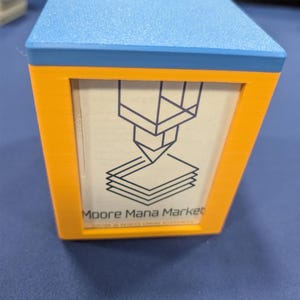 Könnte beinhalten: Eine quadratische, 3D-gedruckte Aufbewahrungsbox mit einem leuchtend orangefarbenen Rahmen und einem hellblauen Deckel. Die Vorderseite zeigt einen weißen Einsatz mit einer dunkelblauen Strichzeichnung einer 3D-Druckerdüse und dem Text Moore Mana Market.