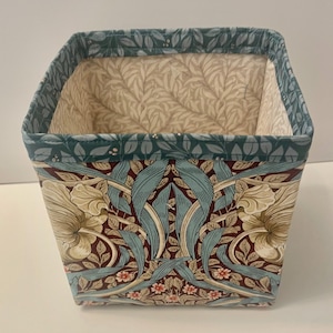 May include: A square fabric storage bin featuring a dark red, light blue, and cream floral pattern on the exterior. The top rim is trimmed in a teal fabric with a leaf print, and the interior is lined with a cream-colored quilted fabric.