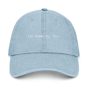 May include: A light blue denim baseball cap with a curved brim and six-panel construction. The front of the cap features the embroidered white text "Too Human for This" centered above the brim. The cap has matching stitching and eyelets.