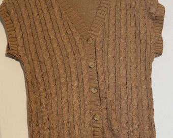 May include: A tan cable-knit sweater vest featuring a V-neckline, short sleeves, and a front button-down closure with five matching tan buttons. The garment is constructed with a vertical cable pattern and ribbed trim at the hem and neckline.