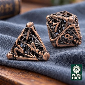 May include: Two hollow, copper-colored metal polyhedral dice with an intricate, open-work filigree design featuring skull motifs. One is a four-sided triangular die and the other is an eight-sided die with visible numbers. A logo reads PURE DICE.