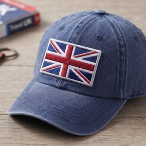 May include: A blue denim baseball cap featuring a rectangular embroidered patch of the Union Jack flag on the front panel. The cap has a curved brim, stitched detailing, and a metal adjustable strap closure on the side for a secure fit.