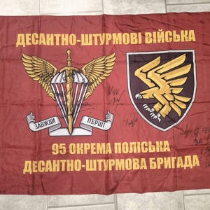 Può includere: Una bandiera rettangolare color marrone con il testo giallo "ДЕСАНТНО-ШТУРМОВІ ВІЙСЬКА" in alto. Presenta un emblema centrale con un paracadute alato e una spada, uno scudo con un'aquila dorata e il testo "95 ОКРЕМА ПОЛІСЬКА ДЕСАНТНО-ШТУРМОВА БРИГАДА" in basso.